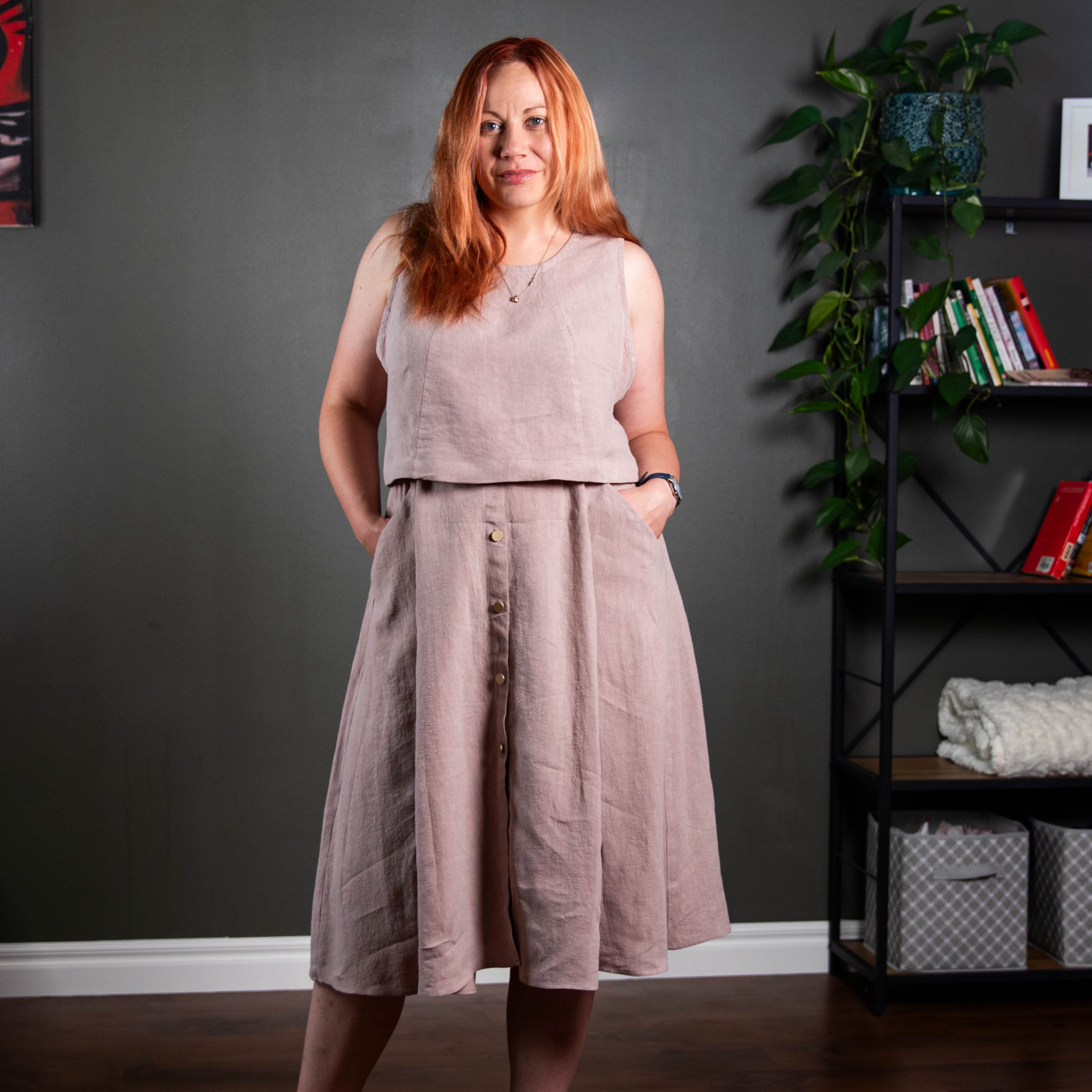 Woman wearing a light pink sleeveless top and wide-leg pants standing in a room with a dark gray wall and a shelf with books and plants.