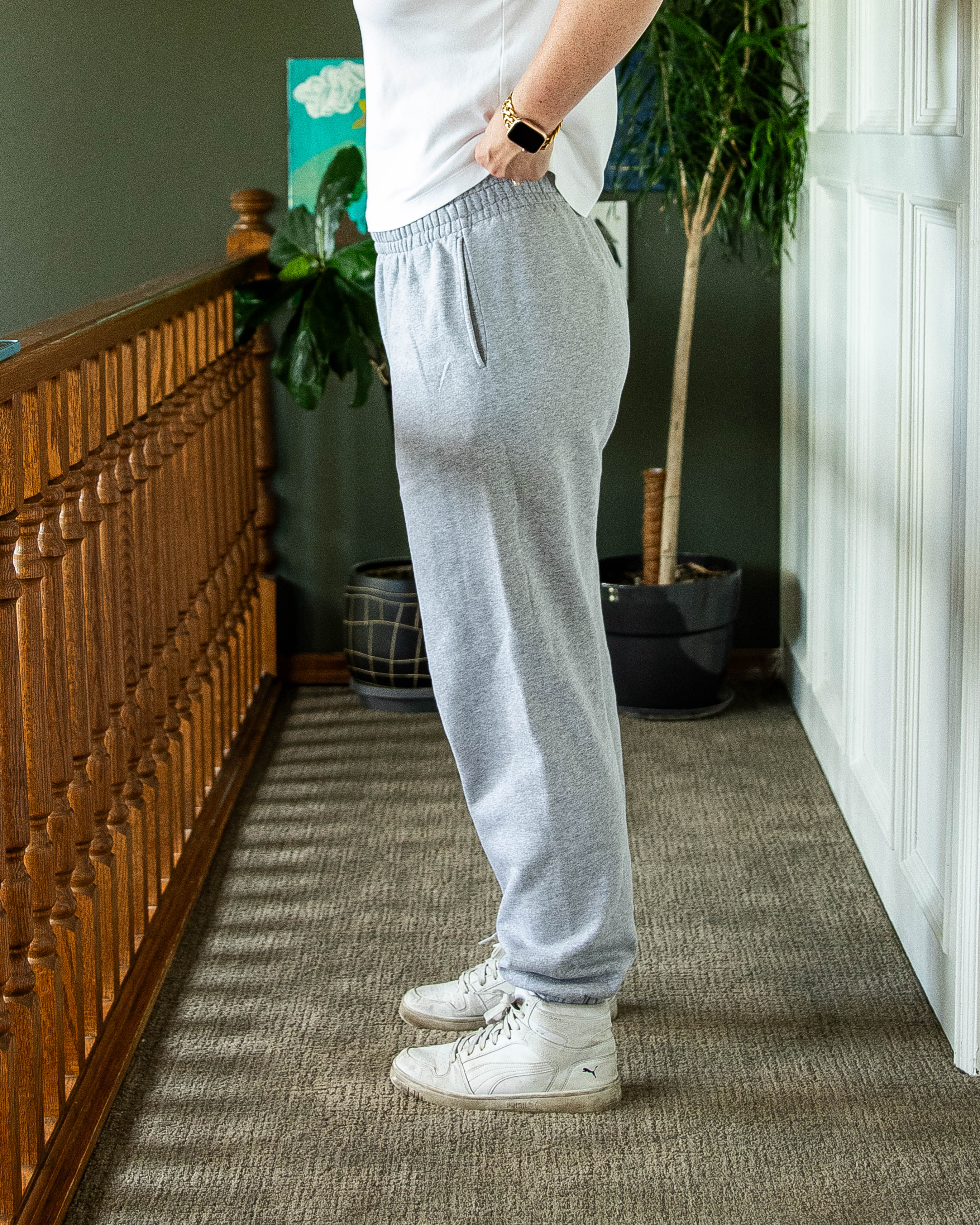 Person, standing side ways, wearing gray sweatpants and white sneakers in a room with a wooden dresser and plants.