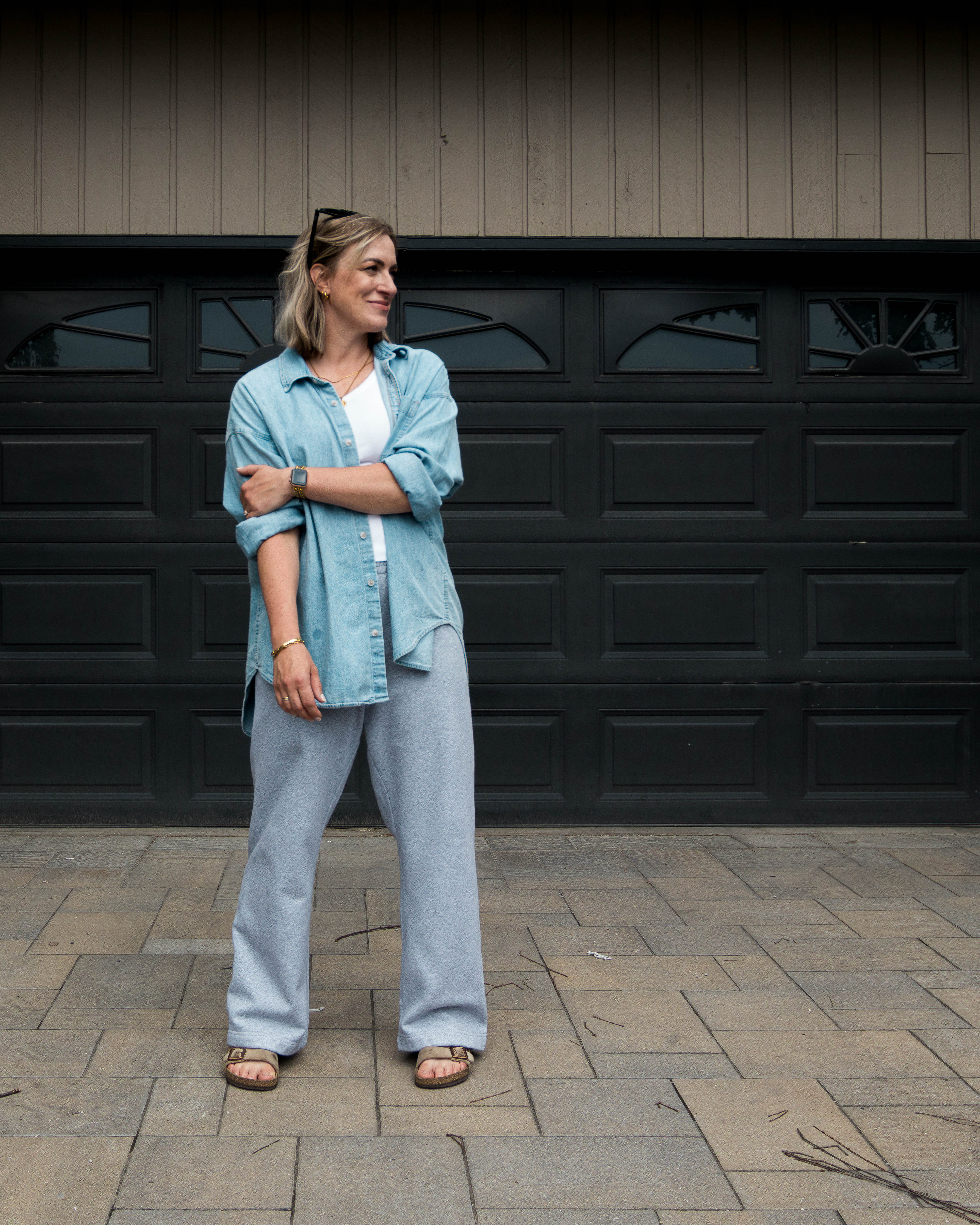Woman standing in front of a black garage door wearing a light blue denim shirt and gray sweat pants.