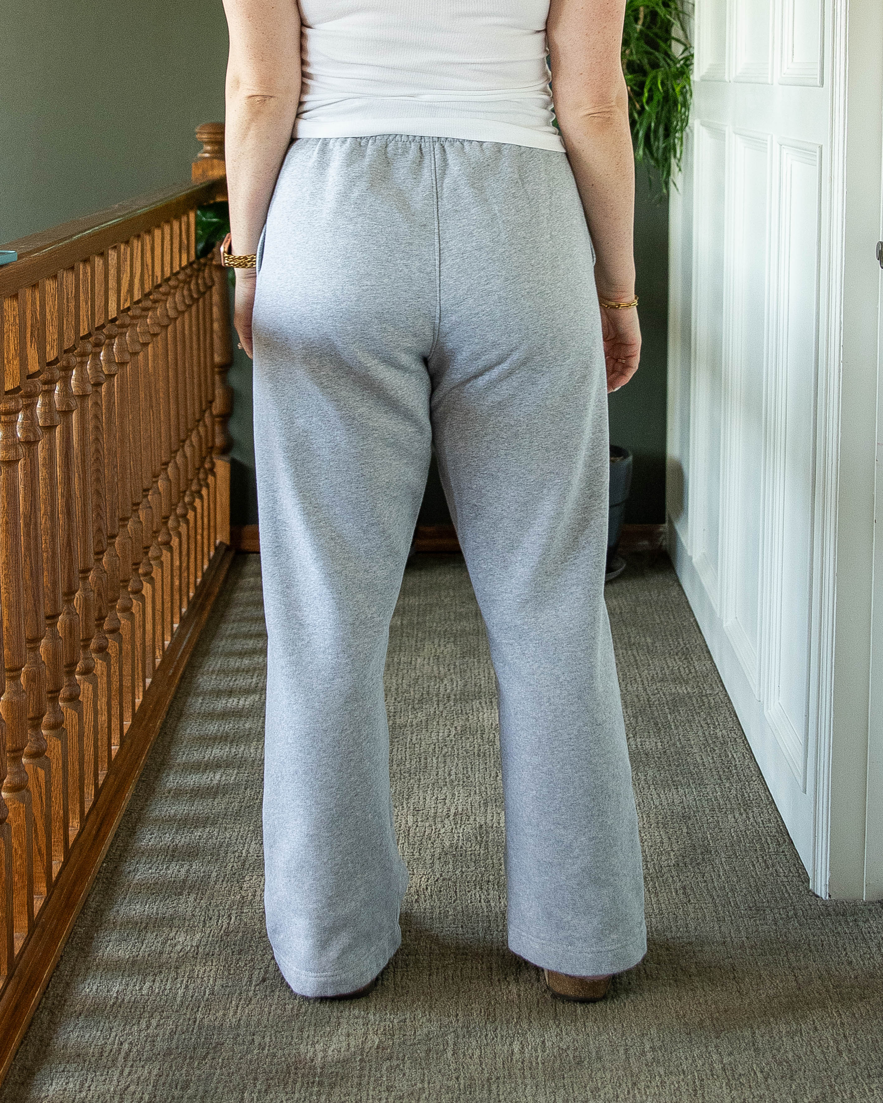 A back view of a person wearing gray sweatpants, waist down. standing in a room with wooden bnnister and a white wall.
