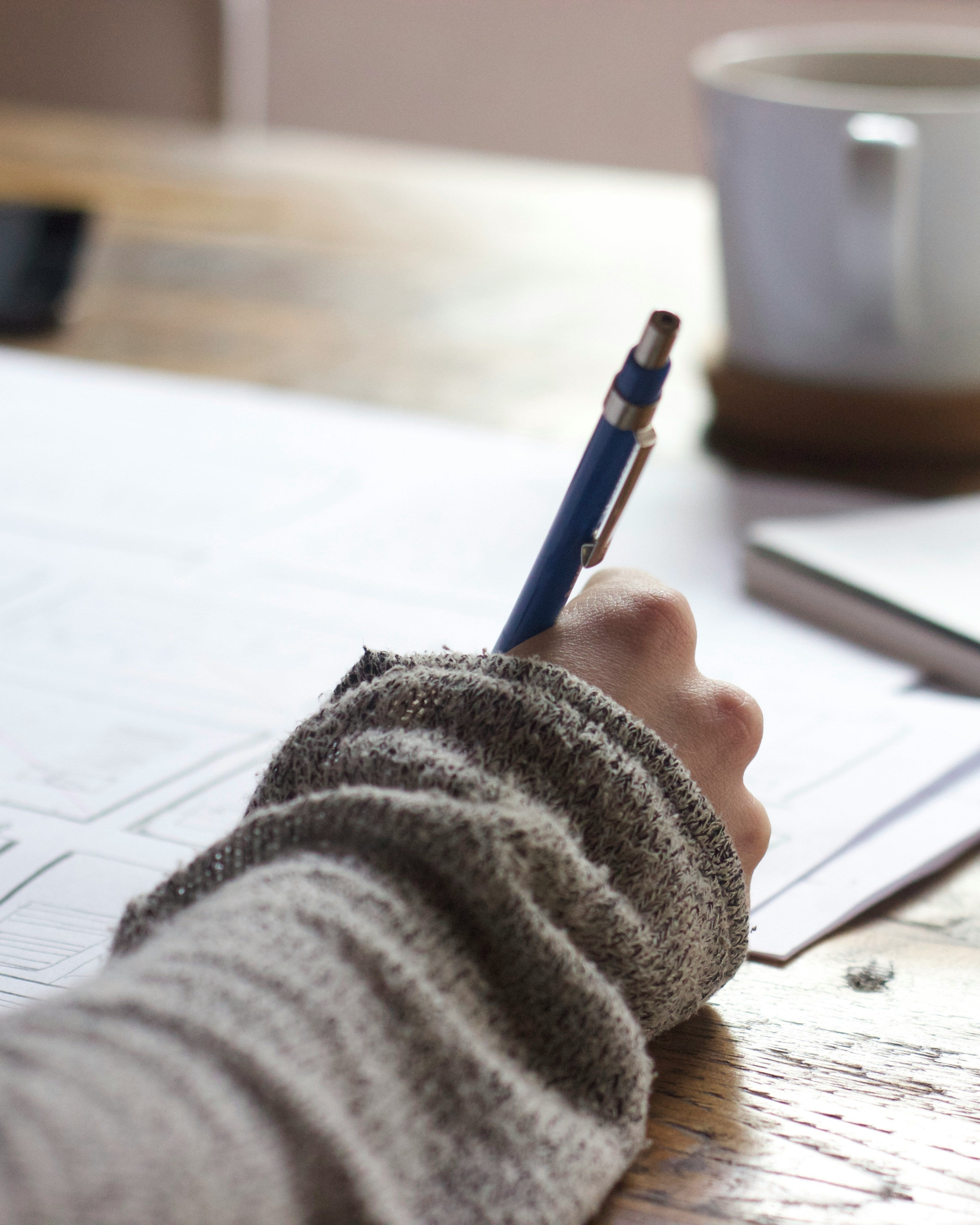 Person writing with a pen on a piece of paper at a desk with a notebook and mug in the background.